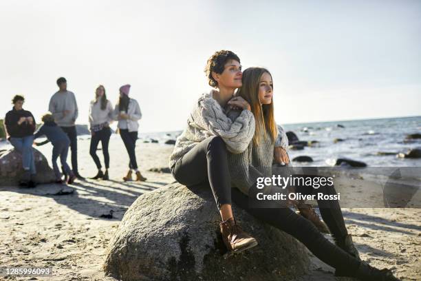 young woman and girl sitting on boulder on the beach with family in background - young teen girl beach foto e immagini stock