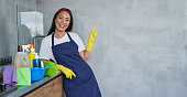 Cheerful young woman, cleaning lady in protective gloves smiling at camera, pointing up while standing in the kitchen with cleaning products and equipment, ready for cleaning the house