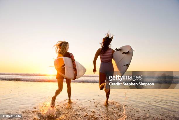 female surfers running on the beach at sunset - california stock-fotos und bilder