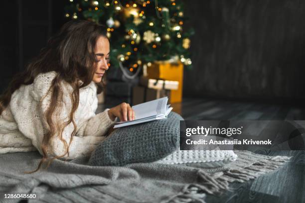 beautiful woman reading book while relaxing on cushions by christmas tree at home. - art books stock-fotos und bilder
