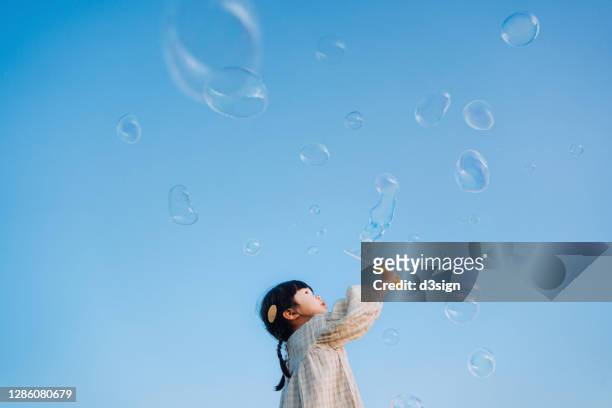 adorable little asian girl holding a bubble wand having fun playing with soap bubbles outdoors in nature park against beautiful blue sky on a lovely day - china games day 1 fotografías e imágenes de stock