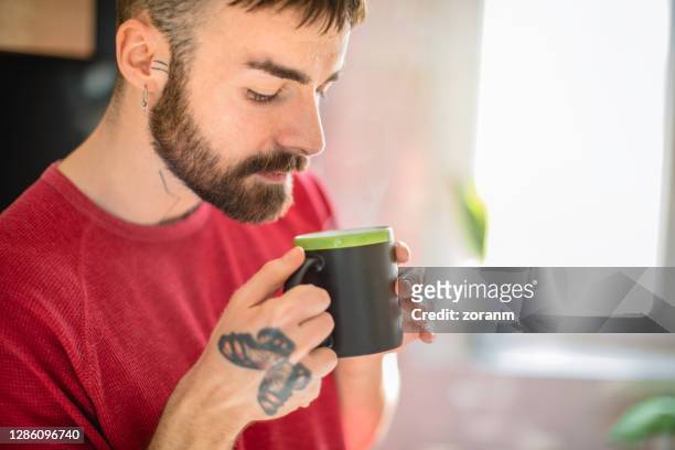 young man with beard holding mug of hot drink and looking at it - butterfly tattoos stock pictures, royalty-free photos & images