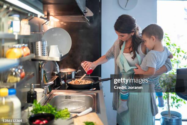 mother with toddler on hip by the stove adding salt to fried meal - sprinkling stock pictures, royalty-free photos & images