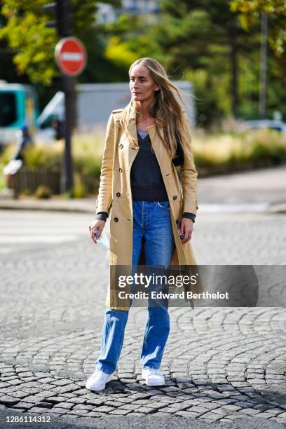 Guest wears a pale yellow long trench coat, a v-neck pullover, blue denim jeans, white sneakers, outside Hermes, during Paris Fashion Week -...