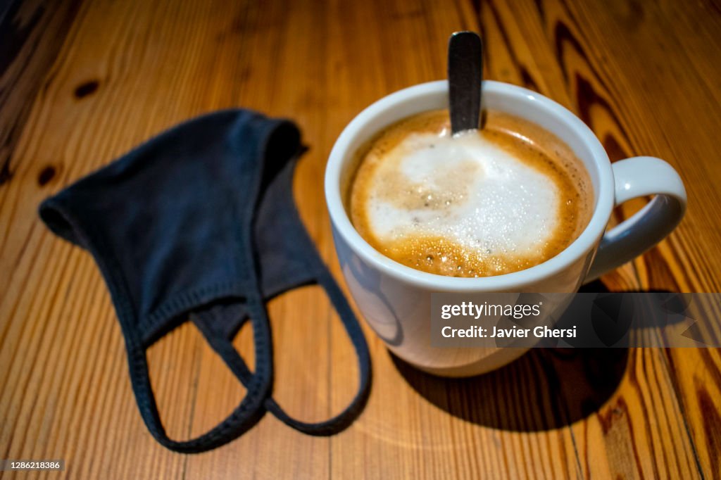 Taza de café con leche y tapaboca (barbijo) sobre mesa de madera