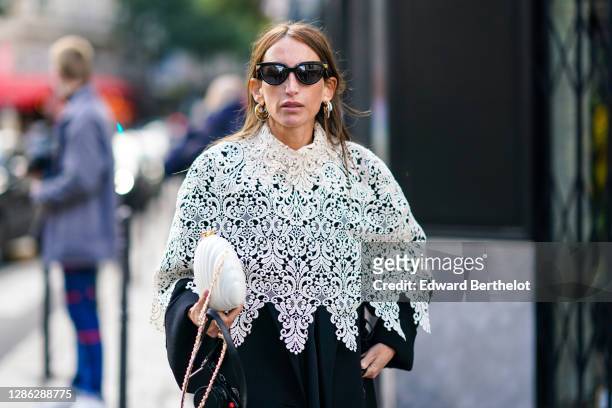 Guest wears sunglasses, earrings, a white top with floral embroidery, a balck long coat, a white bag in the form of a sea-shell, outside Paco...