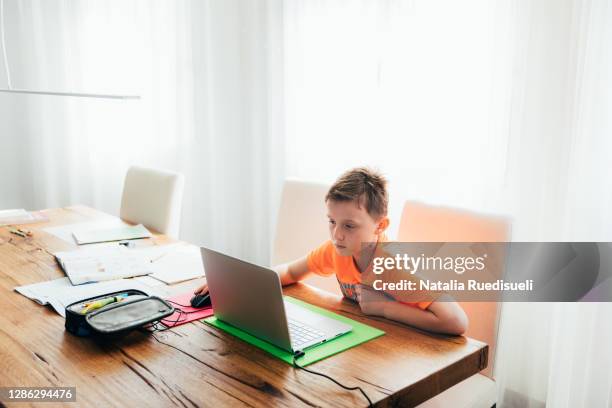 10 years old boy sitting at the dining table at home and doing homeschool on his laptop. - 10 11 years fotografías e imágenes de stock