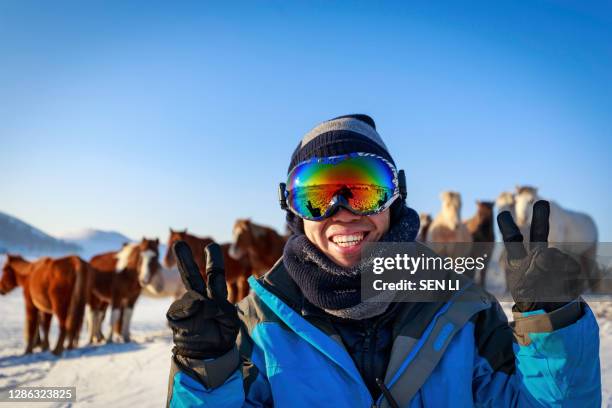 an asian man in winter jacket and ski glasses, smiling happily at the camera, on the snow field in a horse pasture - 中国 stock pictures, royalty-free photos & images