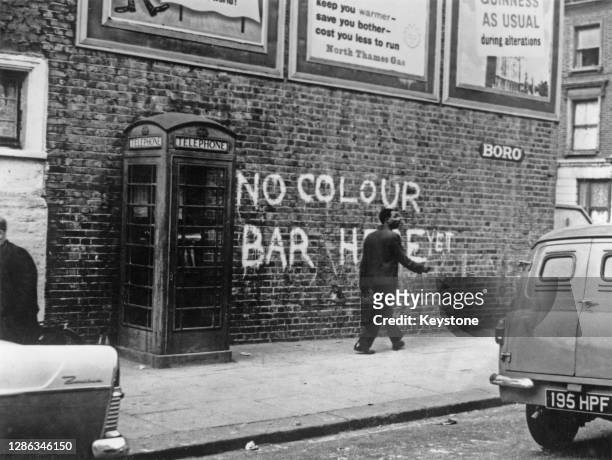 Man walks beside a brick wall onto which is painted graffiti reading 'No colour bar here yet', alongside a telephone box, in Notting Hill, west...