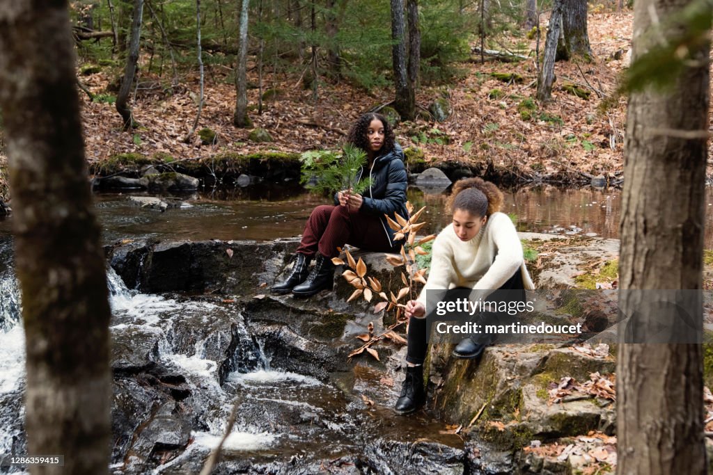 Mixed-race sisters relaxing on river side in autumn nature.