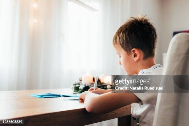 a 9 years old boy sitting at the wooden table and writing christmas greeting cards. cozy ambience of a decorated house with advent wreath and window lights. - 8 9 years stock pictures, royalty-free photos & images