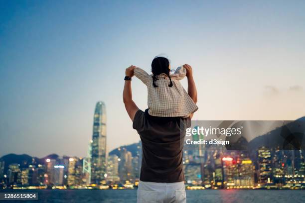 loving young asian father carrying little daughter on shoulders, looking over illuminated urban city skyline against the promenade of victoria harbour at sunset. enjoying father and daughter bonding time together - erwartung stock-fotos und bilder