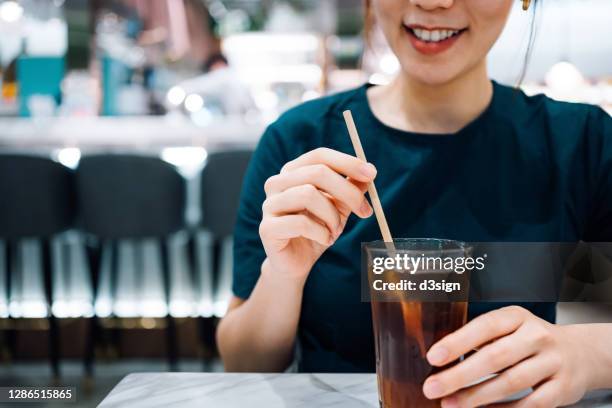 close up of cheerful young asian woman relaxing in a cafe, enjoying a glass of iced tea using an environmentally friendly paper straw. sustainable lifestyle, ecosystem, recycled material concept - rietje stockfoto's en -beelden