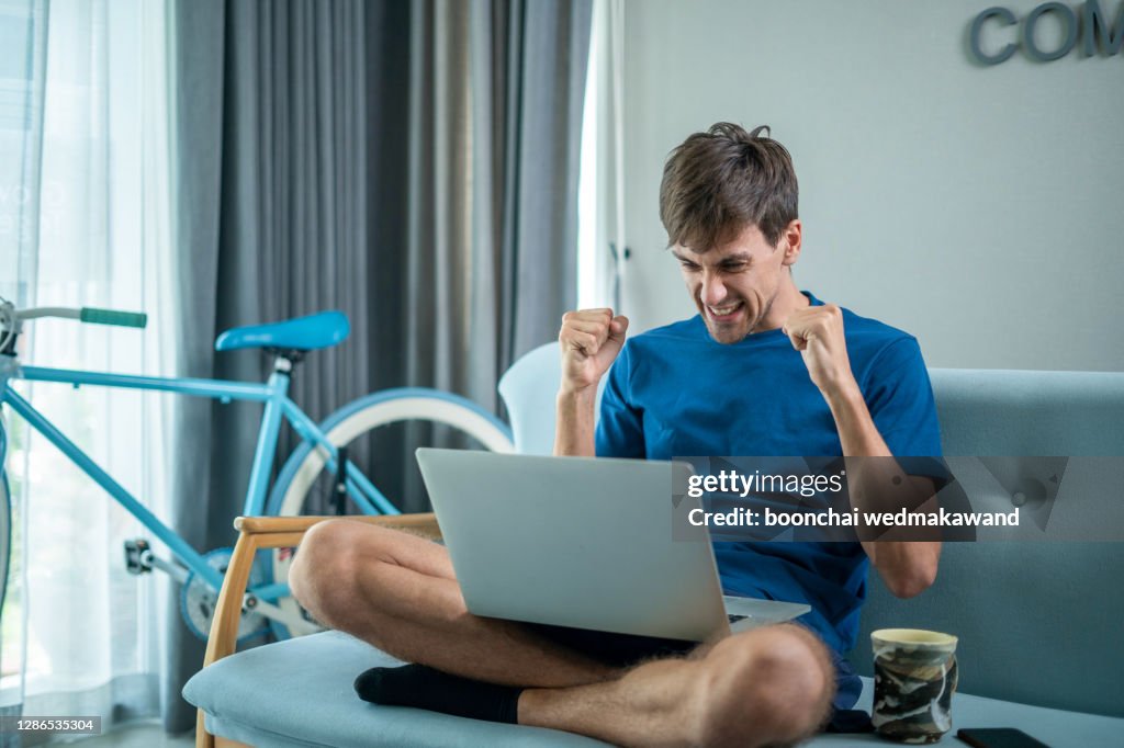 Excited millennial man in glasses sit at table in kitchen feel euphoric win online lottery on laptop, overjoyed young male in spectacles look at computer screen triumph get good news on email