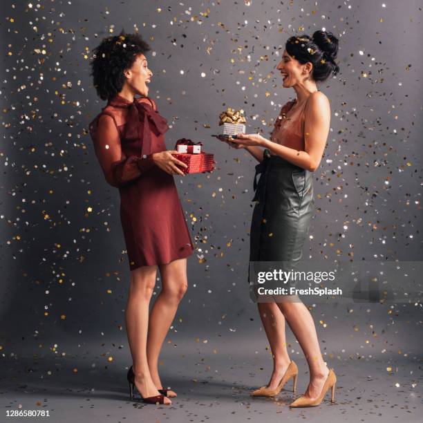 celebration time: two elegant women exchanging christmas gifts at a party, confetti flying all around them - gift exchange stock pictures, royalty-free photos & images