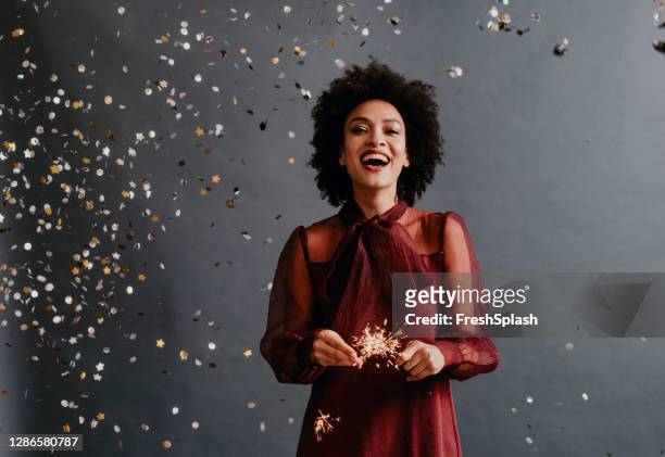 portrait of a smiling african american woman in a red festive dress holding ignited sparklers under confetti - sparklers stock pictures, royalty-free photos & images