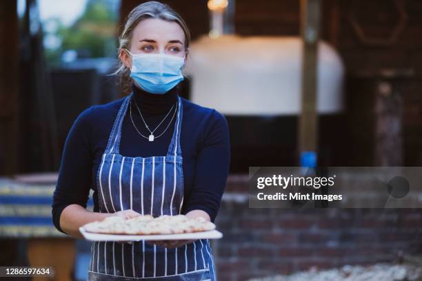 woman waitress in apron and face mask holding fresh pizza on a board - coronavirus restaurant stock pictures, royalty-free photos & images