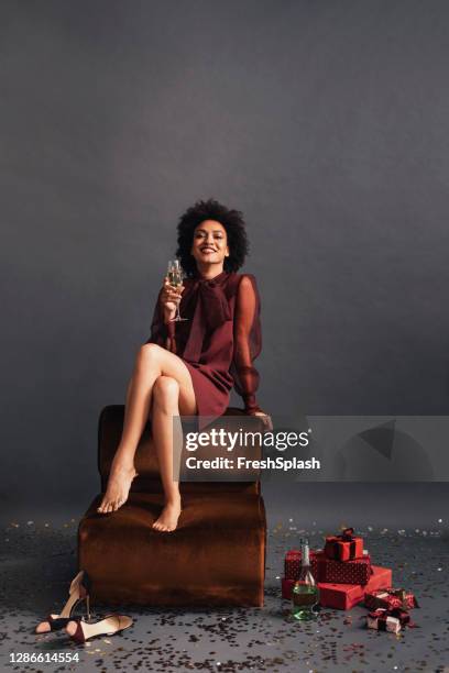 studio shot av en leende afroamerikansk kvinna sitter barfota och med ett glas champagne med en hög med julklappar på golvet - black dress party bildbanksfoton och bilder