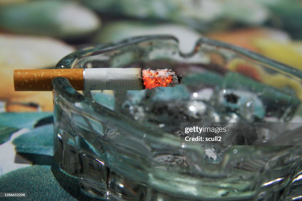 Glass ashtray with a burning half smoking cigarette on the table