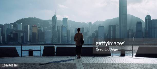 silhouette of young asian woman relaxing by the promenade of victoria harbour overlooking the city, enjoying the spectacular hong kong city skyline during the day - look back at d day stock pictures, royalty-free photos & images