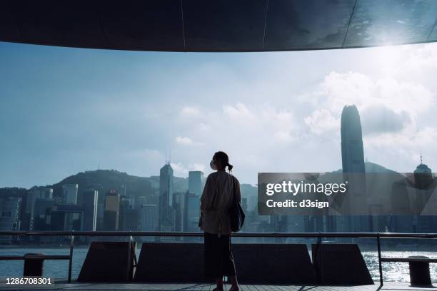 silhouette of young asian woman with protective face mask standing by the promenade of victoria harbour against spectacular hong kong city skyline overlooking the city. in hopes that the epidemic will end soon - end of life care stock pictures, royalty-free photos & images