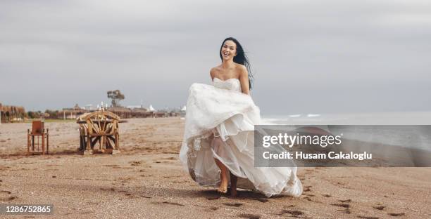 happy beautiful bride is running barefoot in her wedding dress on the beach. - bride running stock pictures, royalty-free photos & images