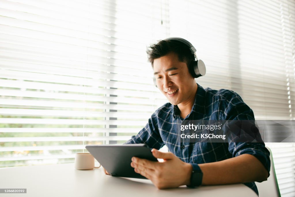 An Asian man relaxing by the home window and typing a message on his digital tablet