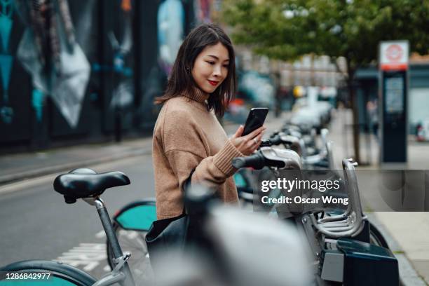 young beautiful woman with smartphone renting bicycle from bike share service in the city - être en mouvement photos et images de collection