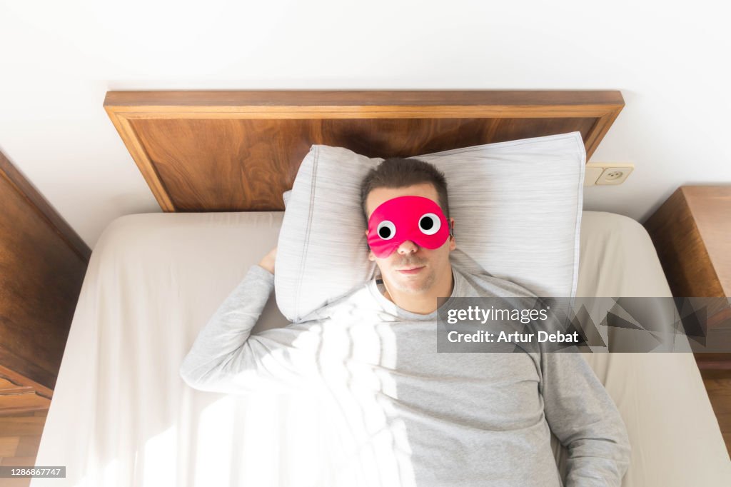 Man wearing funny sleep mask with googly eyes resting in bed during morning.