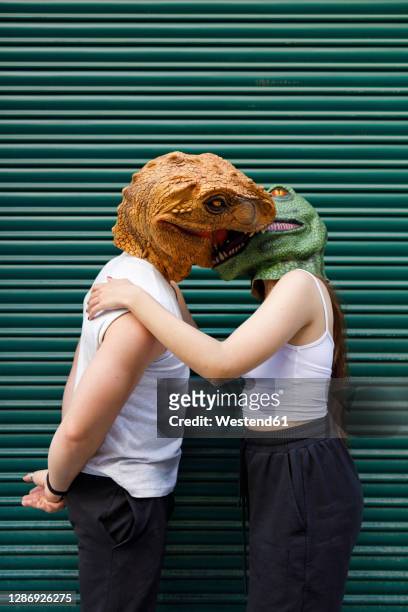 male and female in dinosaur mask kissing against green shutter - casual male standing stock-fotos und bilder