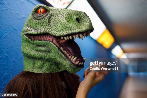 woman applying lipstick while wearing dinosaur mask against blue wall - animal representation stock pictures, royalty-free photos & images