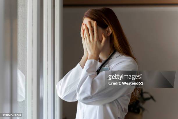 emotionally stressed young female doctor with head in hands standing by window - tristeza imagens e fotografias de stock