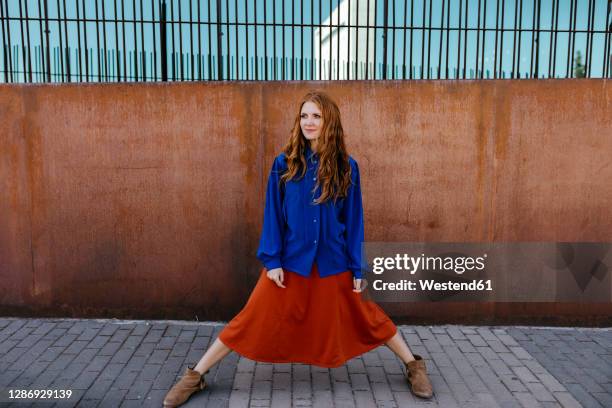 young woman standing with legs apart on footpath against brown wall - legs spread fotografías e imágenes de stock