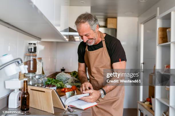 happy mature man reading book in kitchen at home - kochbuch stock-fotos und bilder