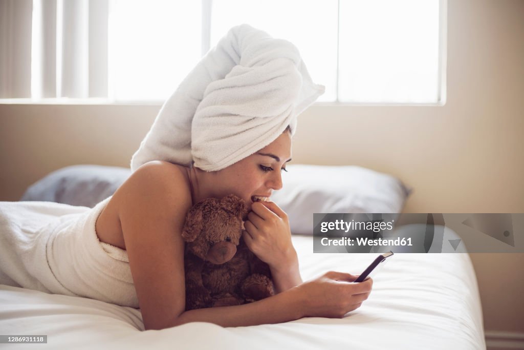 Young woman wrapped in towel using smart phone while lying on bed at home