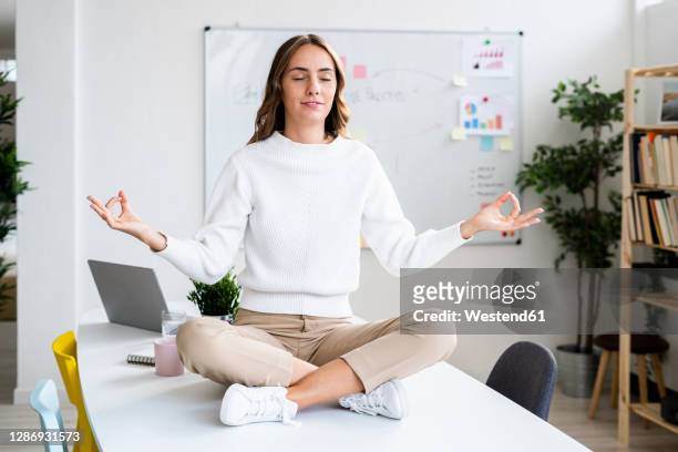 young businesswoman practicing yoga while sitting on table at office - schneidersitz stock-fotos und bilder