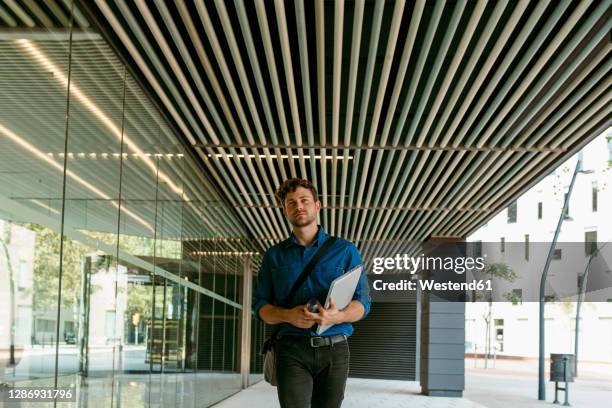 young businessman walking with laptop on walkway at modern office building - effet miroir homme photos et images de collection