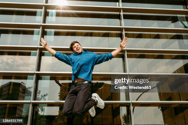 carefree young businessman jumping against modern office building - mann freudensprung sonne vorderansicht leger stock-fotos und bilder