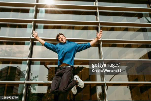 carefree young businessman jumping against modern office building - denim shirt stock pictures, royalty-free photos & images
