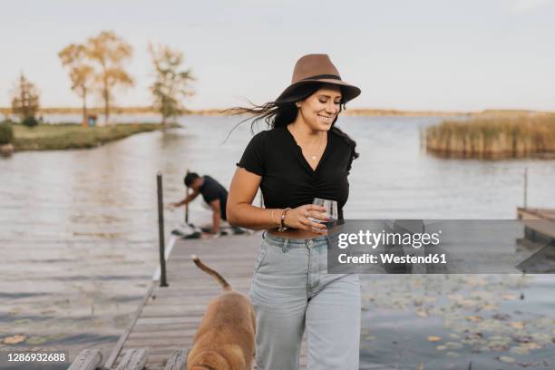 smiling woman holding wineglass while walking with dog and man in background on pier - canada wine stock pictures, royalty-free photos & images