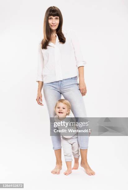 daughter standing in between of mother's leg against white background in studio - baby studio stock-fotos und bilder