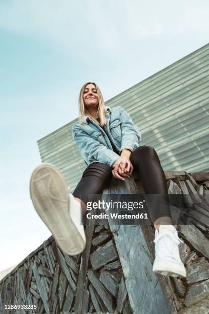 smiling young woman sitting at edge of retaining wall against sky - low angle view stockfoto's en -beelden