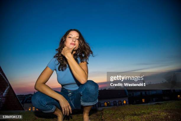 attractive brunette woman with hand on chin crouching against sky during sunset - cool attitude stockfoto's en -beelden