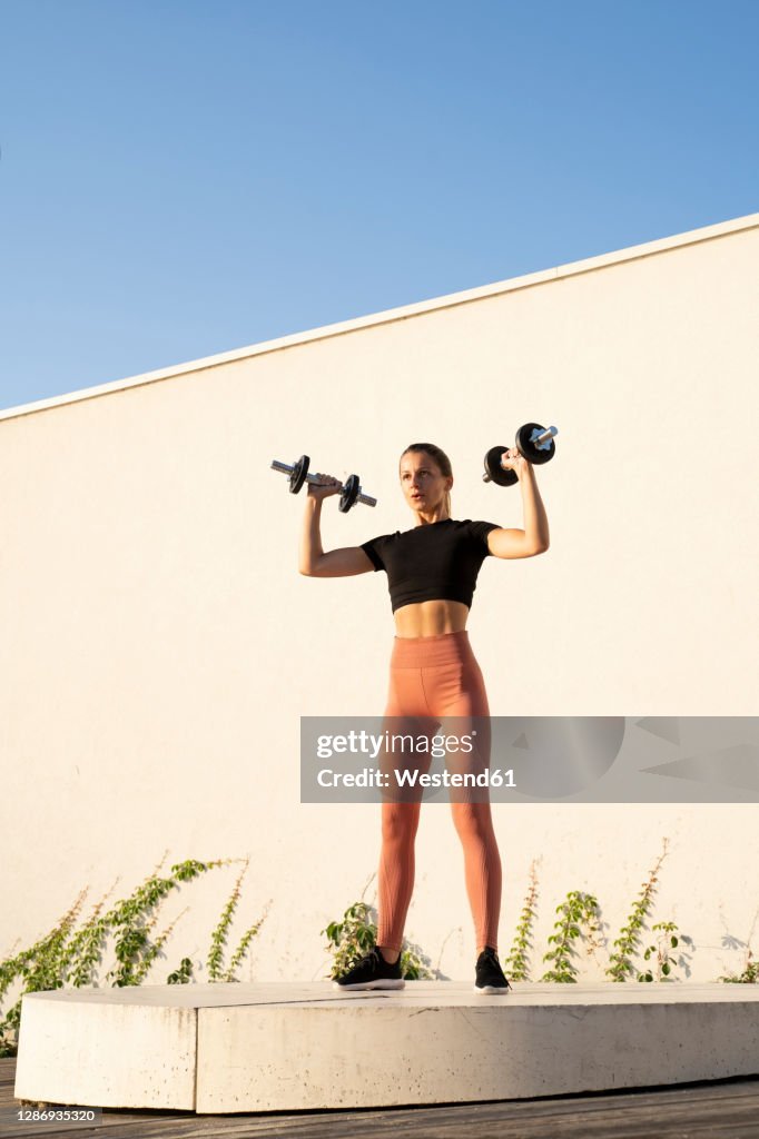 Sportswoman flexing muscle with dumbbell standing on pedestal against wall during sunrise
