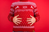 Holidays overeating concept. Close up photo of men in red winter sweater with deers holding his big tummy isolated on red background