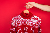 Ugly Christmas sweater party concept. Close up photo of men holding hanger with red winter sweater with deers  isolated on red background