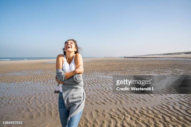 happy young woman hugging self while standing at beach against clear sky - hugging self stock-fotos und bilder