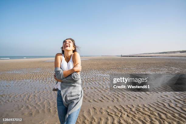 happy young woman hugging self while standing at beach against clear sky - zealand fotografías e imágenes de stock