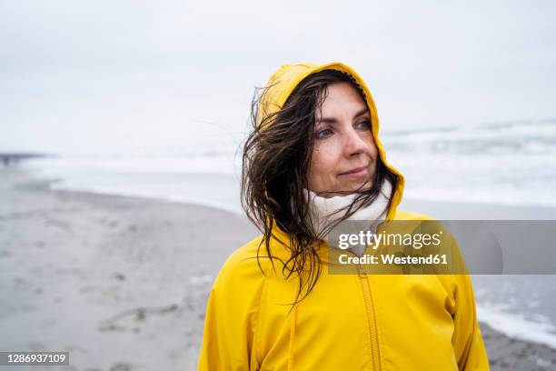 thoughtful mature woman in yellow raincoat standing at beach against sky - kapuze stock-fotos und bilder