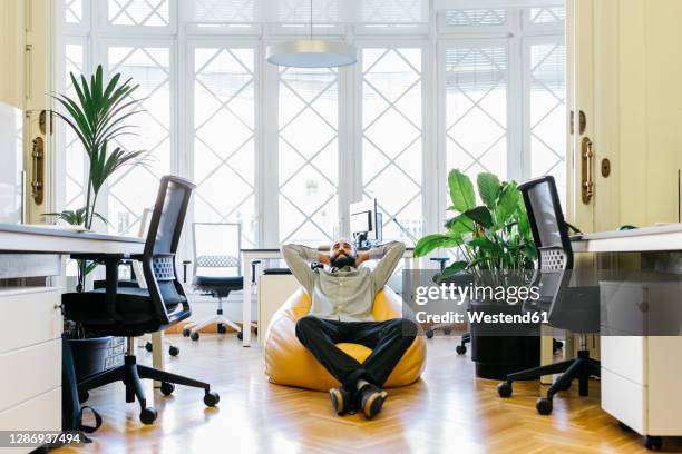 smiling businessman with hands behind head relaxing on bean bag at workplace - beanbag chair stockfoto's en -beelden